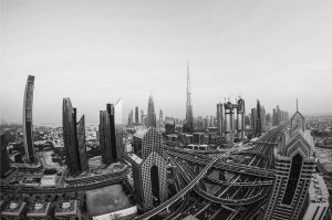 Aerial view of the Dubai skyline along Sheikh Zayed Road in black and white, with the Burj Khalifa, construction cranes and a multi-level interchange.
